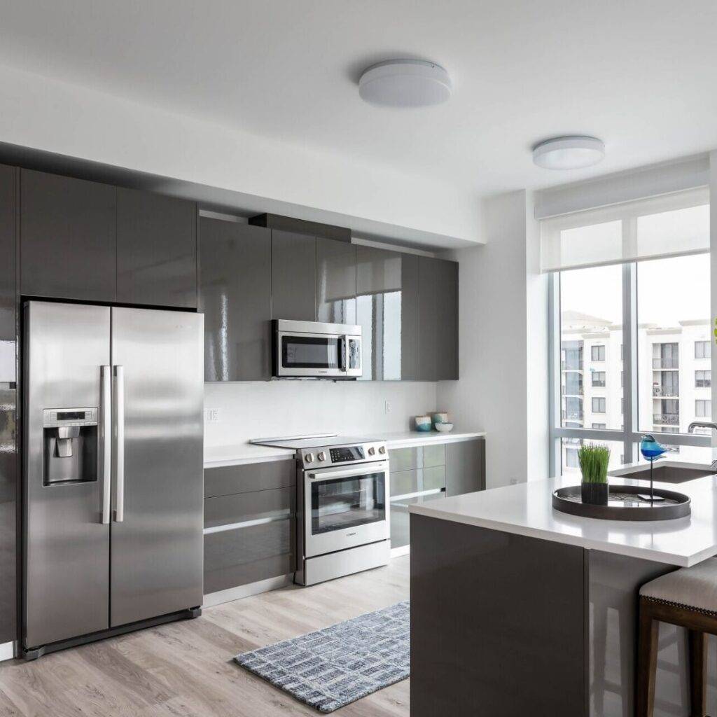 Modern kitchen with sleek gray cabinets, stainless steel appliances, white countertops, and a large window.