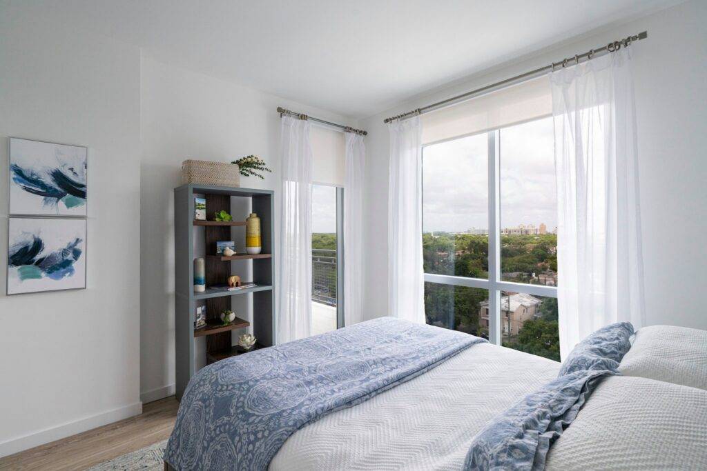 Modern bedroom with large windows, white walls, cozy bed, and a stylish bookshelf.