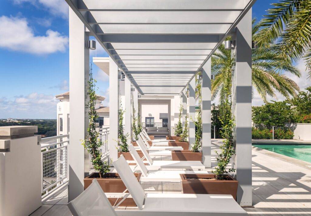 Modern outdoor lounge with white pergola, sun loungers, and a palm tree beside a pool, under a blue sky.