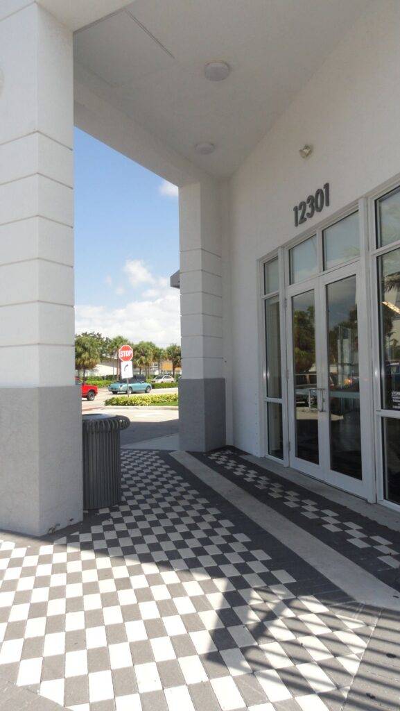 Checkered entrance of a modern building with large glass doors and a sunny backdrop.