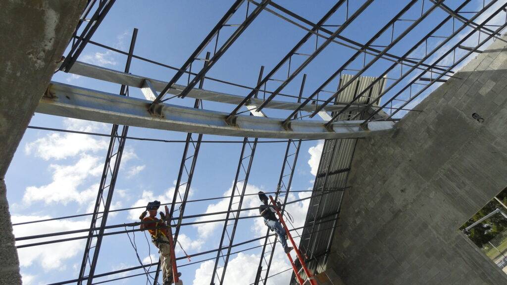 Construction workers on ladders assembling a large metal framework at a building site against a blue sky.