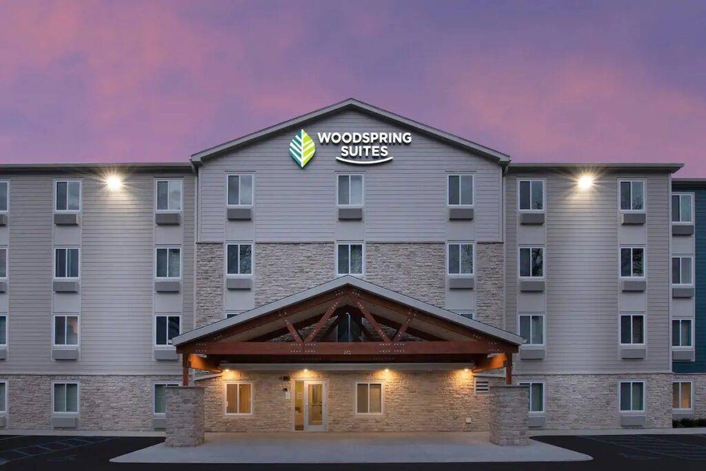 Woodspring Suites exterior at dusk with a lit sign against a pink and purple sky