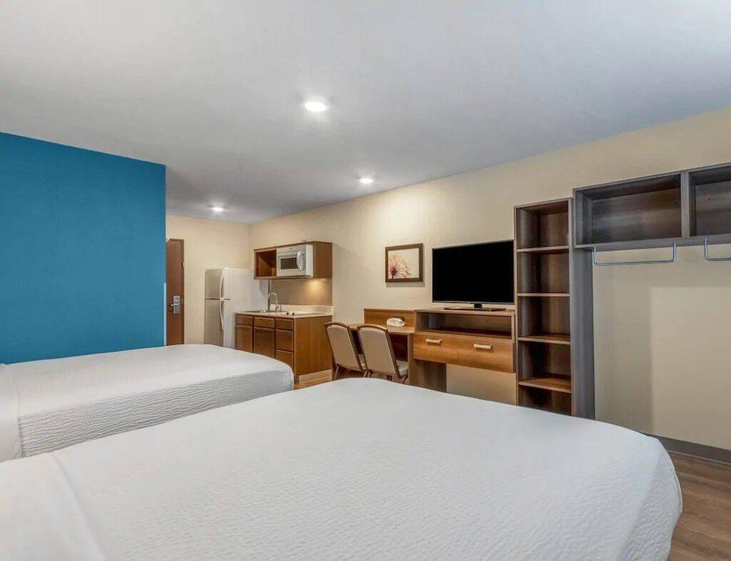 Modern hotel room with two beds, flat-screen TV, kitchenette, and open wardrobe.