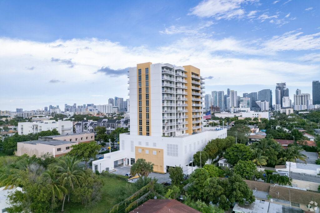 A modern high-rise building amid lush greenery with a cityscape backdrop under a clear blue sky.