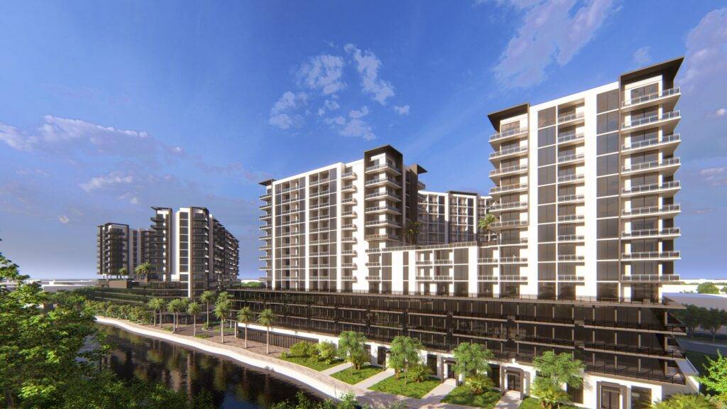 Modern apartment buildings under a clear blue sky by a calm river, surrounded by lush greenery.