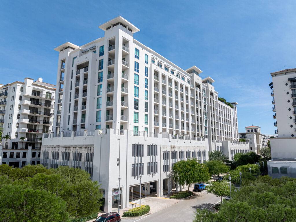 Aerial view of a modern white apartment complex under a clear blue sky, surrounded by green trees.
