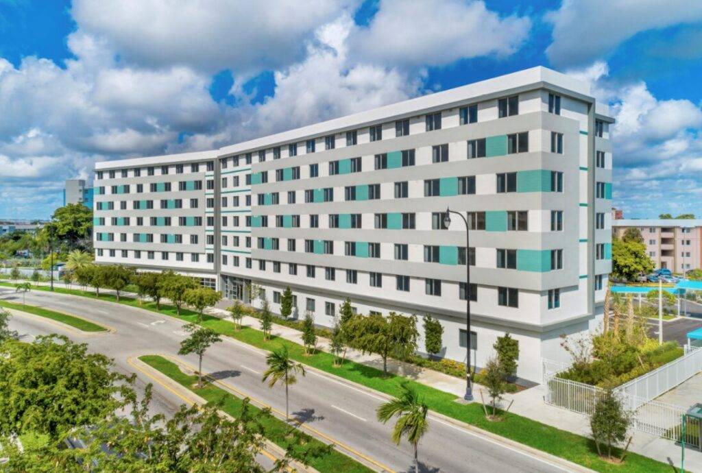 Modern six-story apartment building with a white and teal facade under a partly cloudy sky.