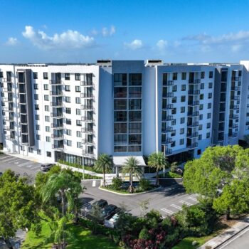 Aerial view of a modern multi-story apartment building surrounded by trees and clear skies.