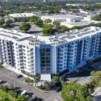 Aerial view of a modern white apartment complex with lush greenery and city surroundings.