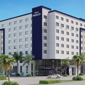 Modern hotel building with palm trees and cars in front, set against a clear blue sky.