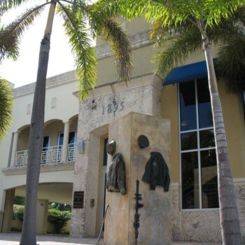 Modern building facade with palm trees and a stone sculpture in front.