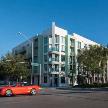 Modern apartment building at a busy urban intersection with a red convertible passing by.