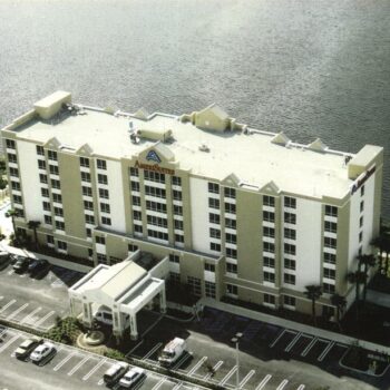 Aerial view of a multi-story hotel building beside a serene lake with parking area in front.