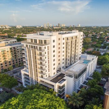 Aerial view of a modern high-rise building surrounded by green trees and cityscape.