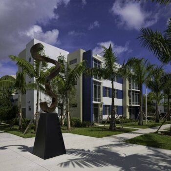 Modern art sculpture outside a contemporary building surrounded by palm trees under a partly cloudy sky.