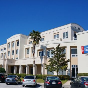 A modern educational building with palm trees and parked cars against a clear blue sky.