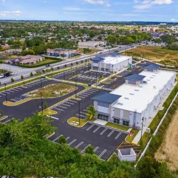 Aerial view of a modern building complex surrounded by a spacious parking area and lush greenery.