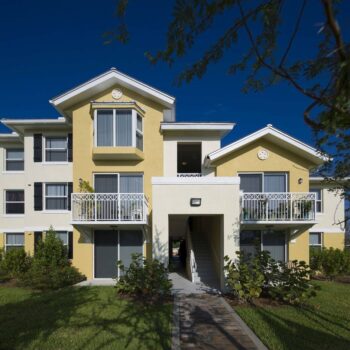 Yellow and white modern apartment building with balconies and lush landscaping.