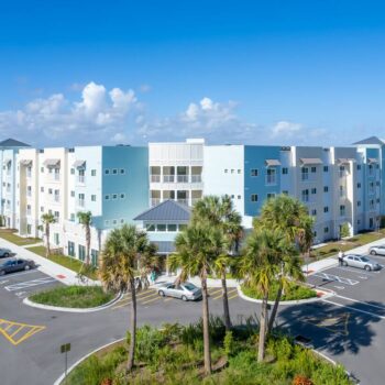 Modern apartment complex with pastel blue facade, palm trees, and a spacious parking area under a clear blue sky.