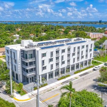 Aerial view of a modern white apartment building surrounded by lush greenery and a distant ocean backdrop.