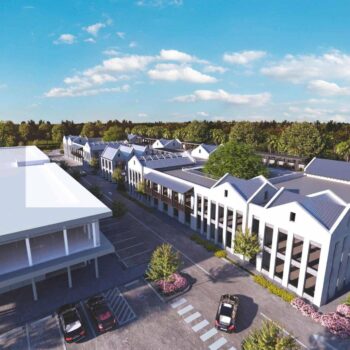 Aerial view of a modern commercial complex with spacious parking, surrounded by lush greenery under a clear blue sky.