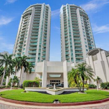 Twin modern high-rise buildings with palm trees and a circular driveway under a bright blue sky.